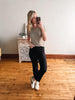 Woman taking a mirror selfie wearing a beige t-shirt and black pants in a room with wooden flooring.