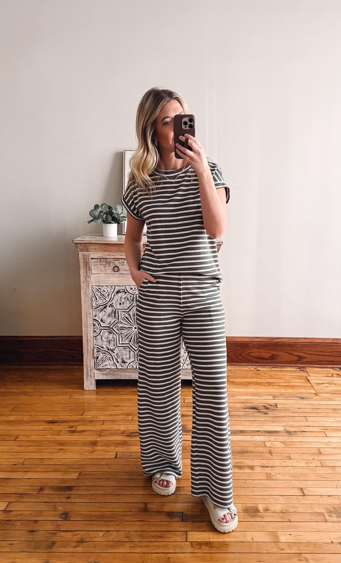 Woman taking a mirror selfie wearing a striped outfit in a room with wooden flooring and a decorative cabinet.