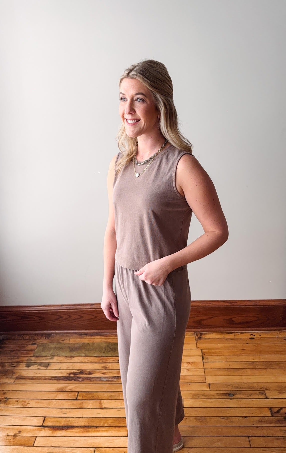 Woman wearing a brown sleeveless top and pants standing against a white wall with wooden flooring.