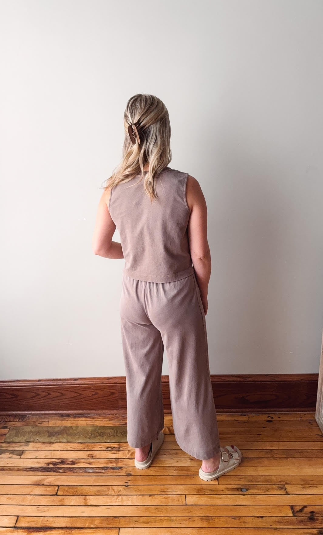 Person wearing a pink sleeveless top and pants standing in a room with wooden flooring and a white wall.