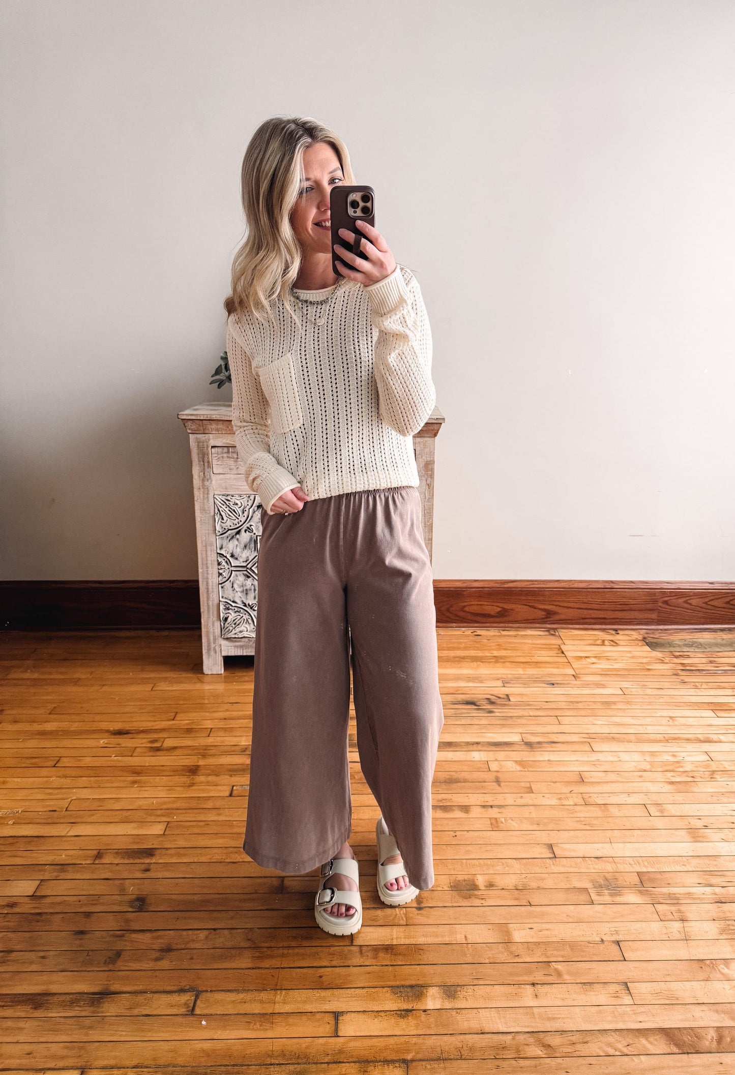 Woman taking a mirror selfie wearing a white sweater and brown pants in a room with wooden flooring.