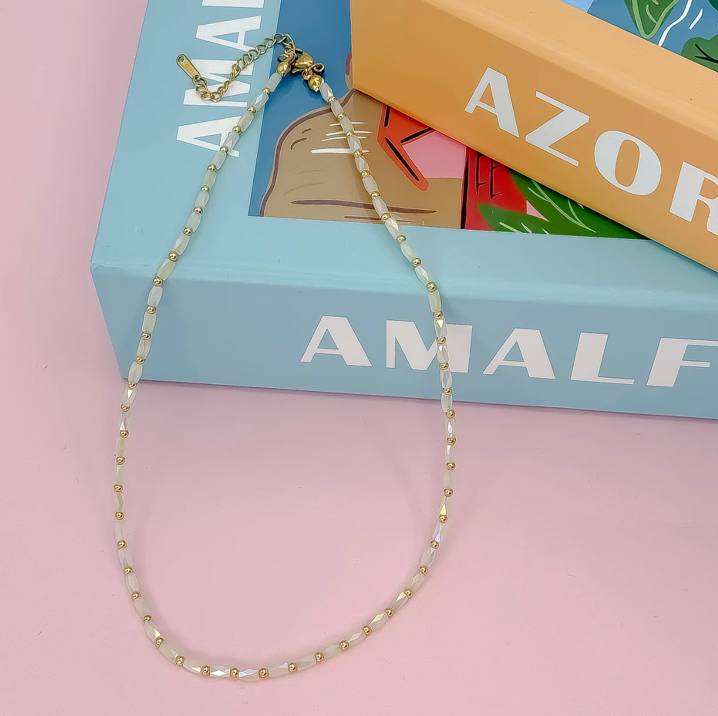 Necklace with gold and white beads on a pink surface with a blue box in the background