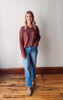 Woman wearing a maroon sweater and blue jeans standing on a wooden floor against a white wall.