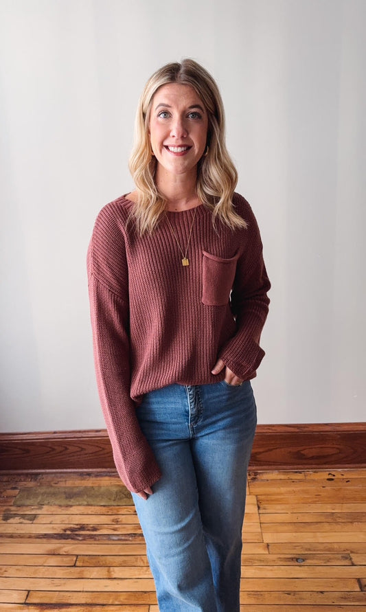Woman wearing a maroon long-sleeve top and blue jeans standing against a plain wall.