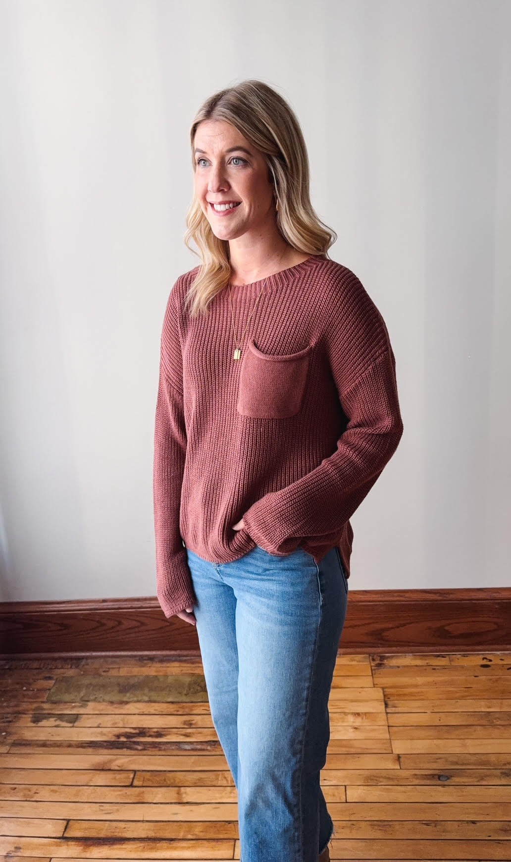 Woman wearing a maroon sweater with a pocket and blue jeans standing in a room with wooden flooring.