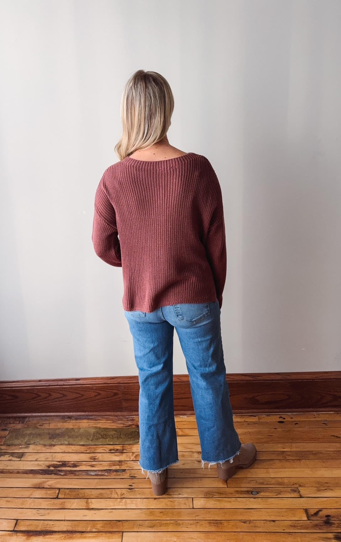 Person wearing a maroon sweater and blue jeans standing on a wooden floor with a white wall background