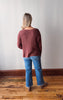 Person wearing a maroon sweater and blue jeans standing on a wooden floor with a white wall background