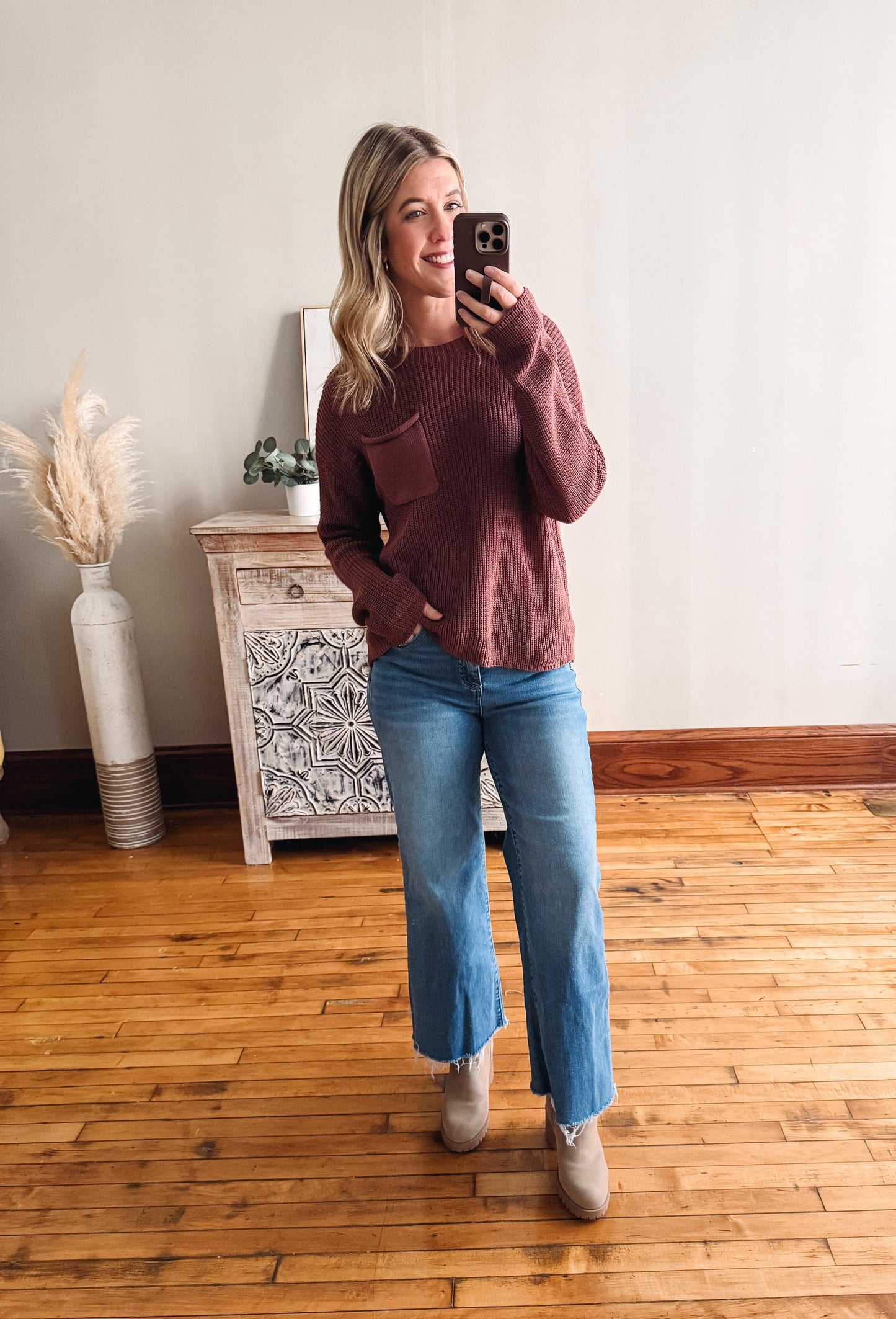 Woman taking a mirror selfie wearing a maroon sweater and blue jeans in a room with wooden flooring.