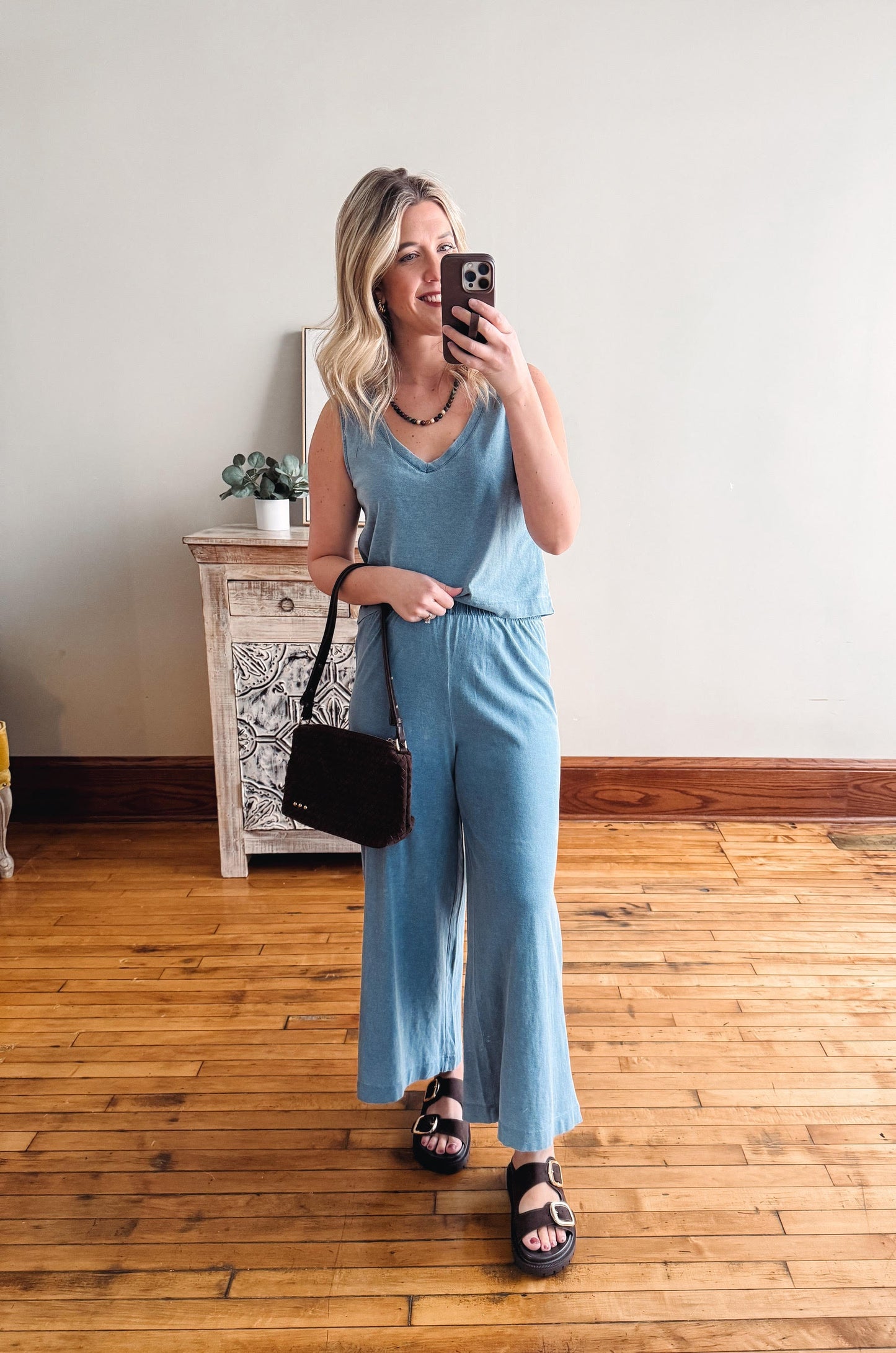 Woman taking a mirror selfie wearing a blue jumpsuit in a room with wooden flooring.