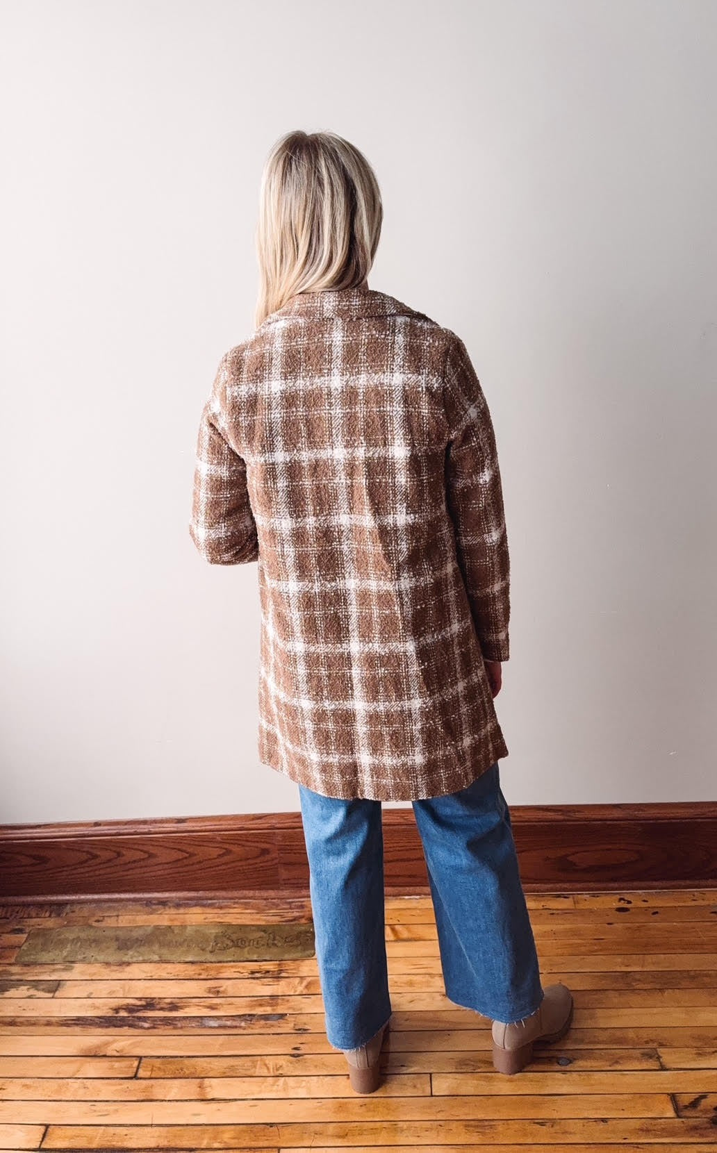 Person wearing a plaid coat and blue jeans standing on a wooden floor against a white wall.