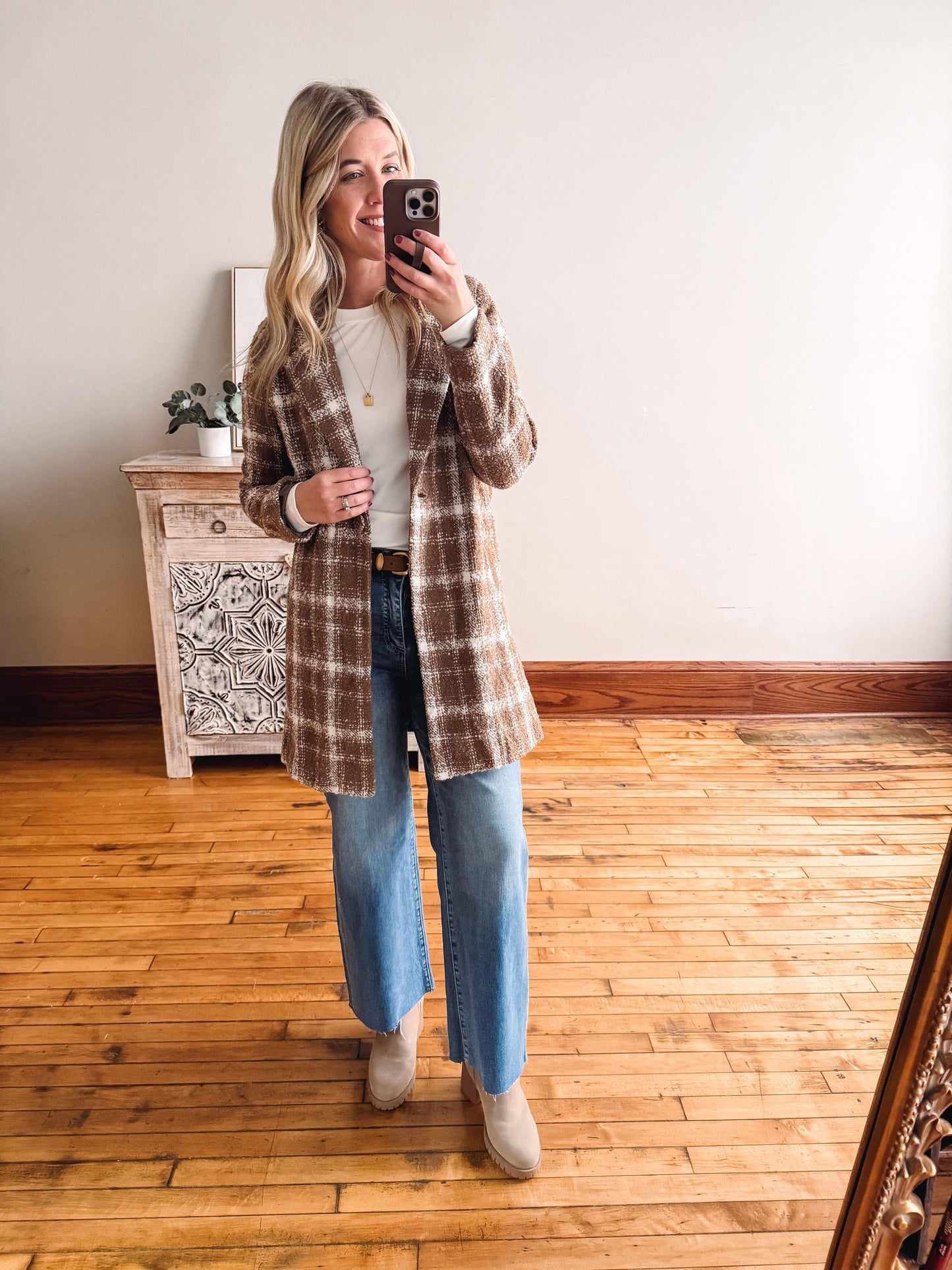 Woman taking a mirror selfie wearing a plaid coat, white shirt, blue jeans, and beige boots in a room with wooden flooring.