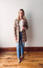 Woman wearing a plaid coat, white shirt, blue jeans, and gray shoes on a wooden floor.