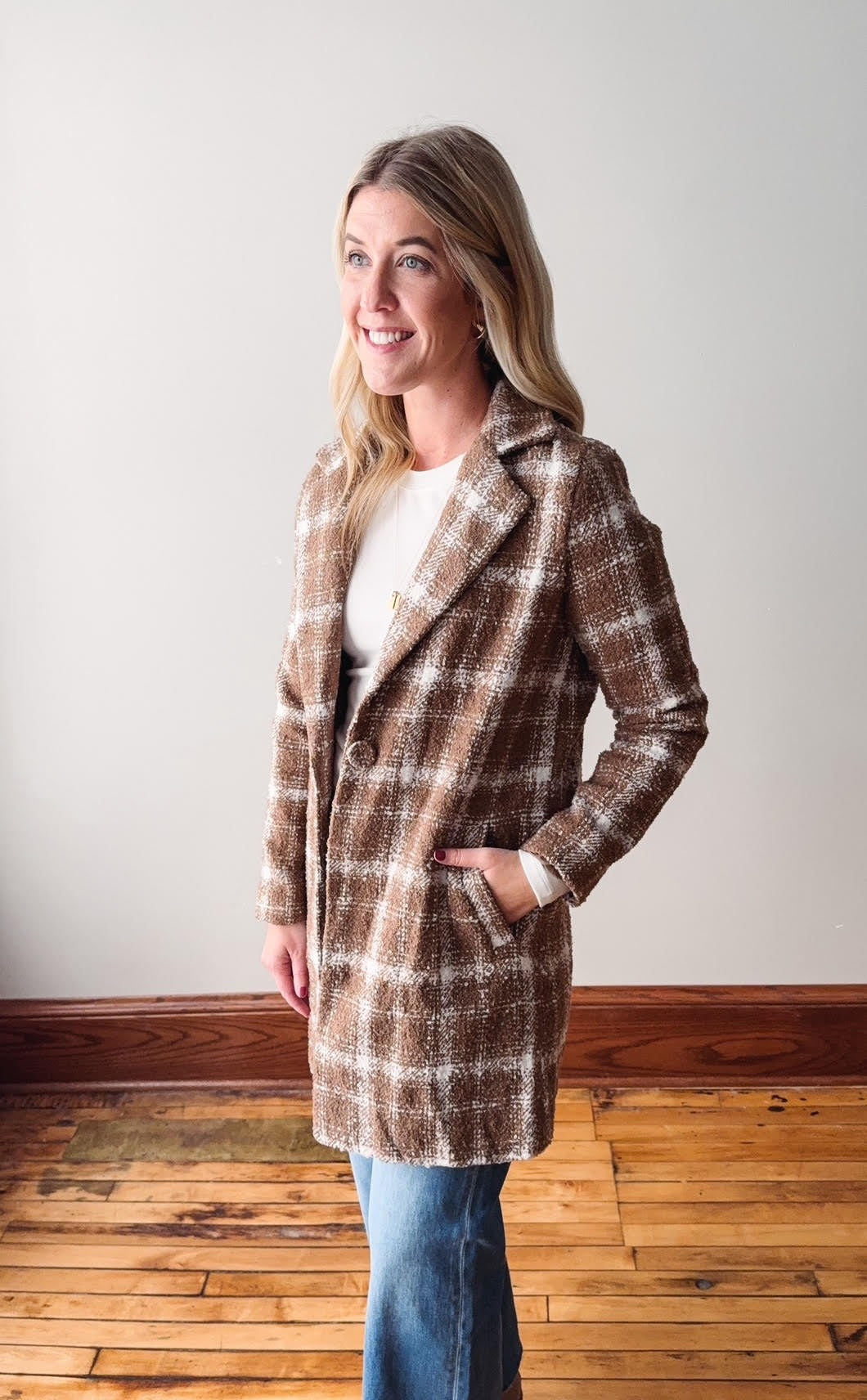 Woman wearing a plaid coat standing in a room with wooden flooring and a white wall.