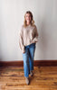 Woman wearing a beige sweater and blue jeans standing on a wooden floor against a white wall.