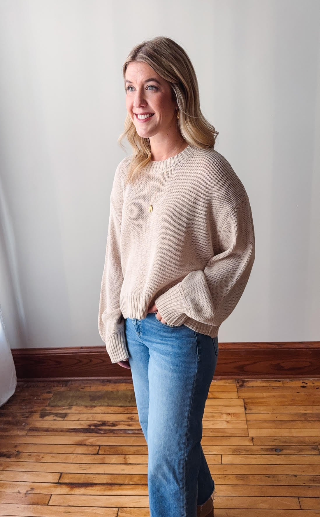 Woman wearing a beige sweater and blue jeans standing in a room with wooden flooring and white walls.