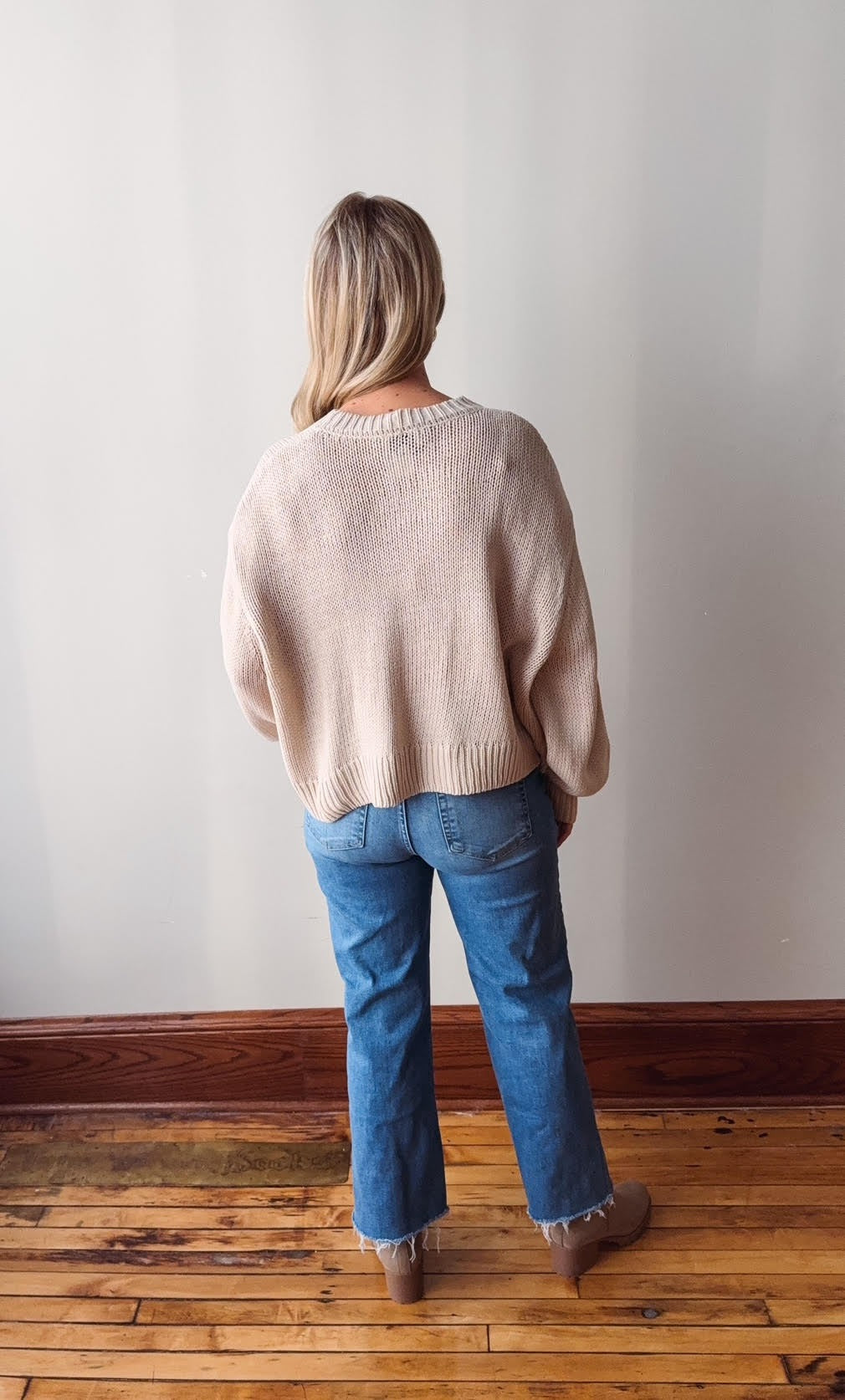 Person wearing a beige sweater and blue jeans standing against a plain white wall.