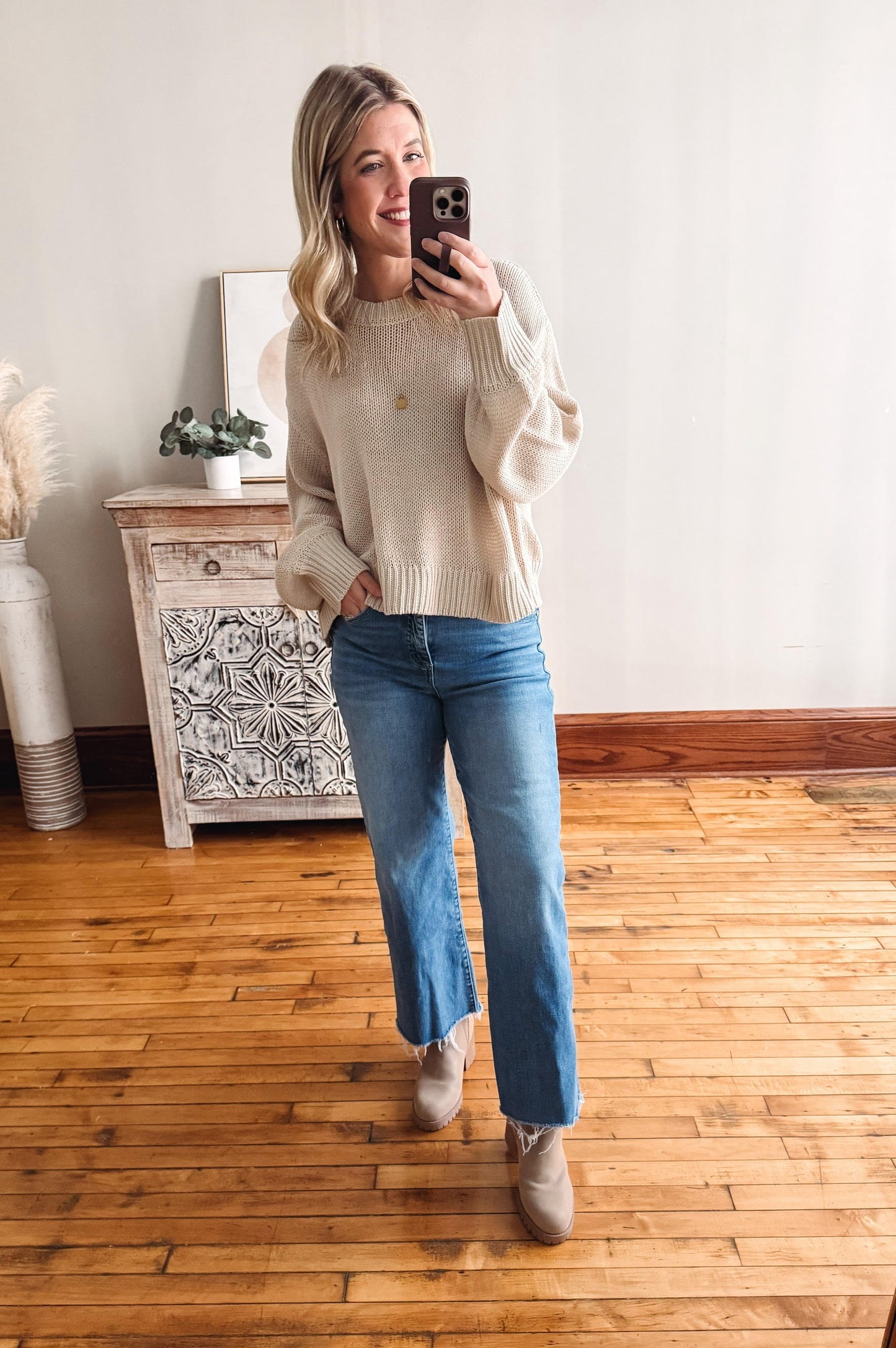 Woman taking a mirror selfie wearing a beige sweater and blue jeans in a room with wooden flooring.
