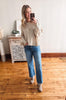 Woman taking a mirror selfie wearing a beige sweater and blue jeans in a room with wooden flooring.