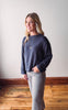 Woman wearing a navy blue sweater and gray pants standing in a room with wooden flooring and white walls.