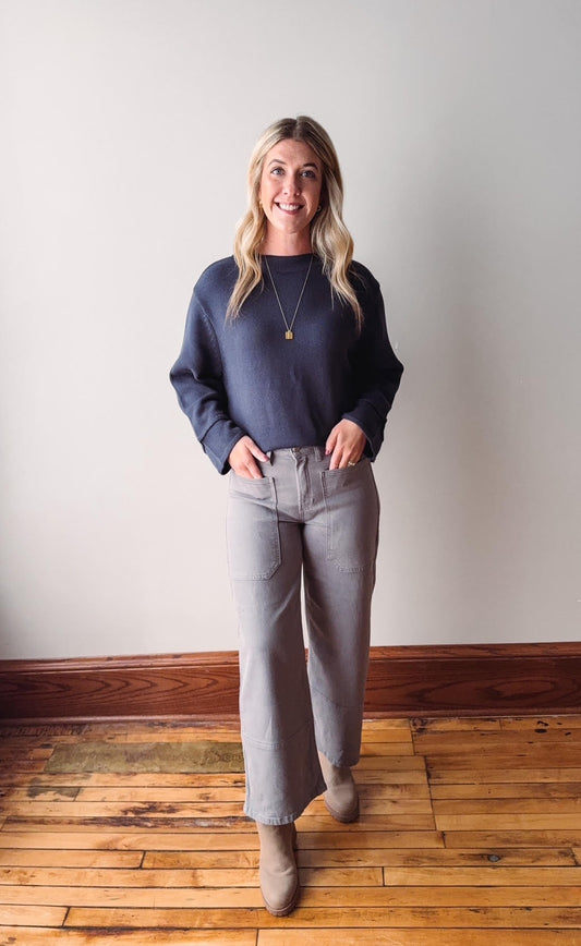 Woman wearing a blue sweater and gray pants standing on a wooden floor with a white wall background