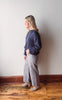 Woman standing against a white wall wearing a navy sweater and light gray pants.