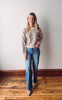 Woman wearing a striped sweater and blue jeans standing on a wooden floor against a white wall.