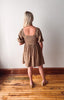 Woman wearing a brown patterned dress standing on a wooden floor with a white wall background