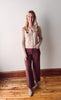 Woman wearing a taupe cardigan with floral buttons and maroon pants standing against a plain wall.