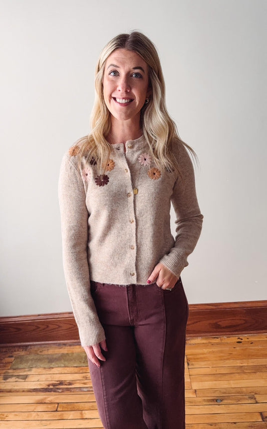 Woman wearing a taupe cardigan with floral embellishments against a plain wall.