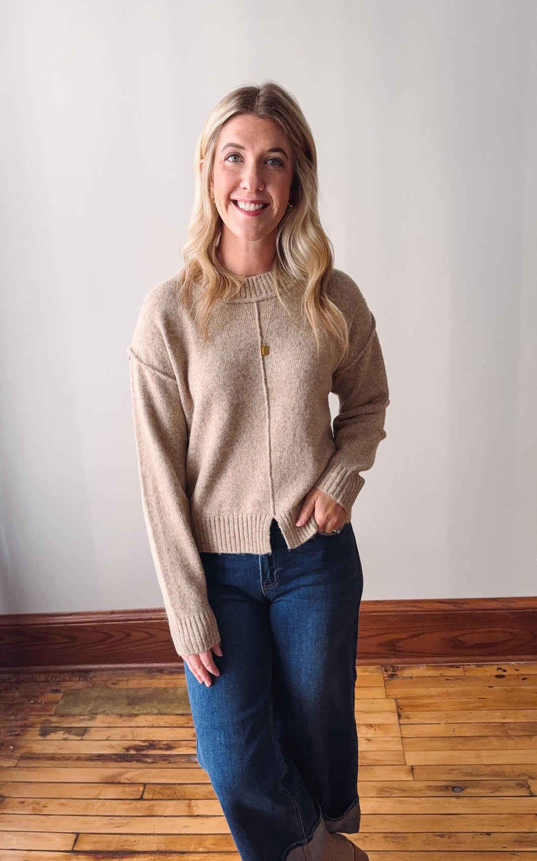 Woman wearing a beige sweater and blue jeans standing in a room with wooden flooring and white walls.