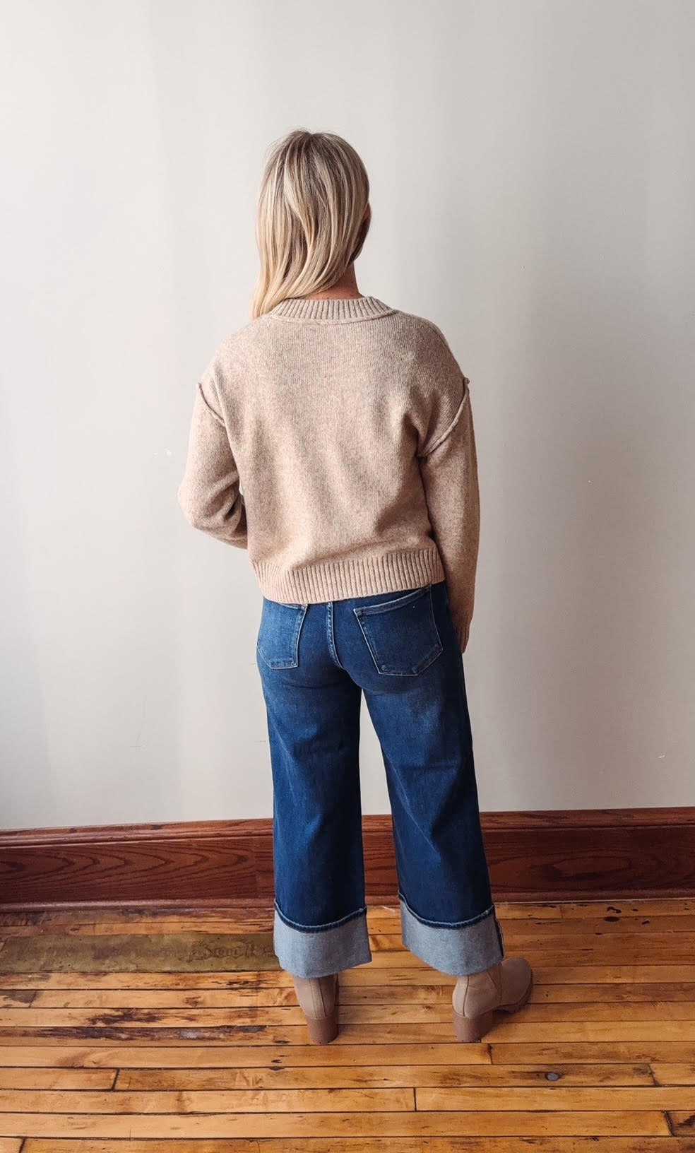 Person wearing a beige sweater and blue jeans standing against a plain wall.