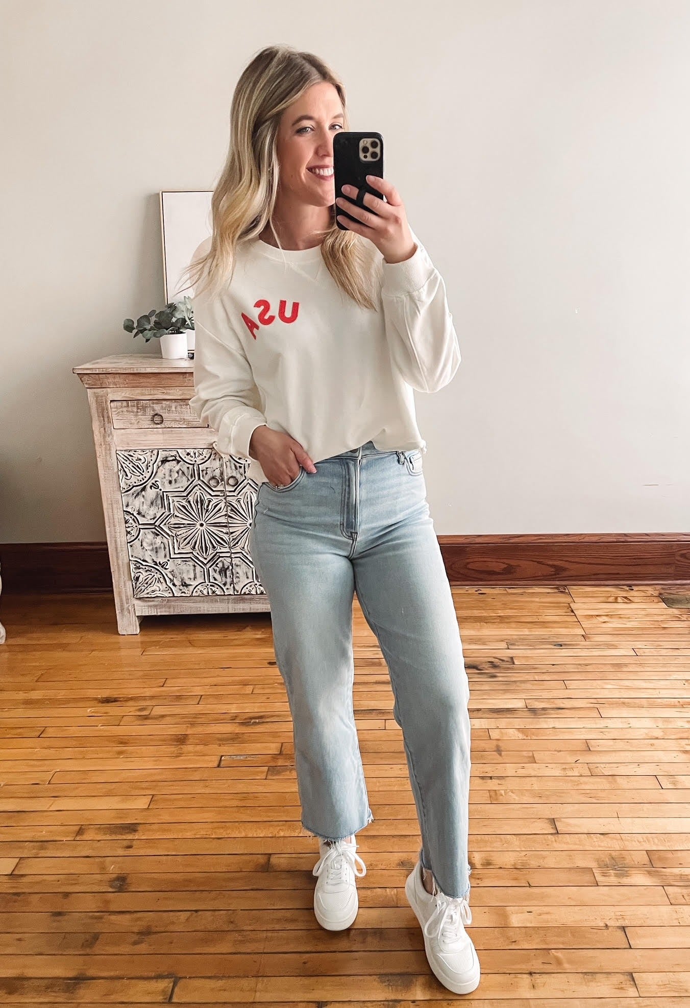 Woman taking a mirror selfie wearing a white sweatshirt and light blue jeans.