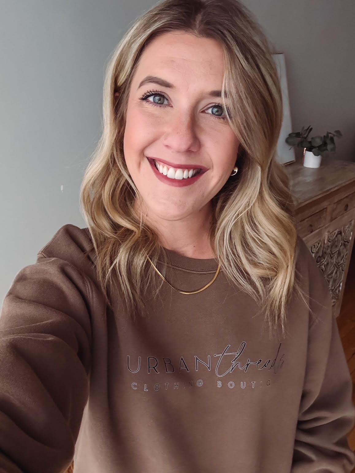 Woman wearing a brown sweatshirt with a logo, smiling indoors.
