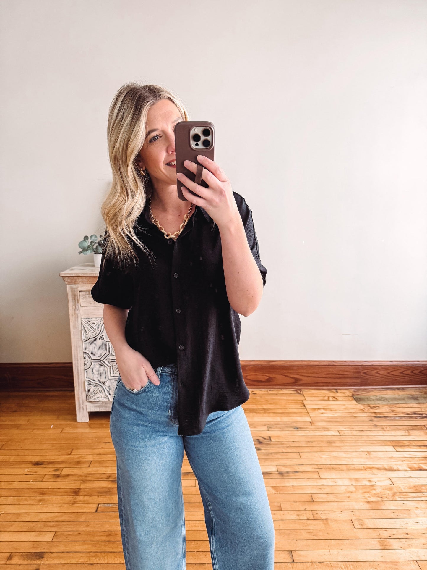Woman taking a mirror selfie wearing a black shirt and blue jeans in a room with wooden flooring.
