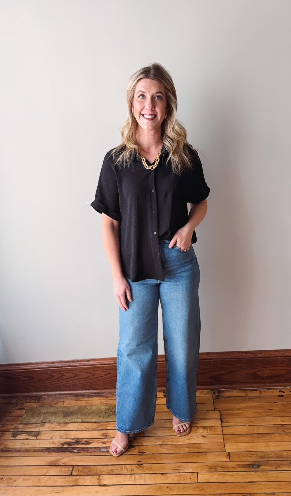 Woman wearing a black top and blue jeans standing against a plain wall.