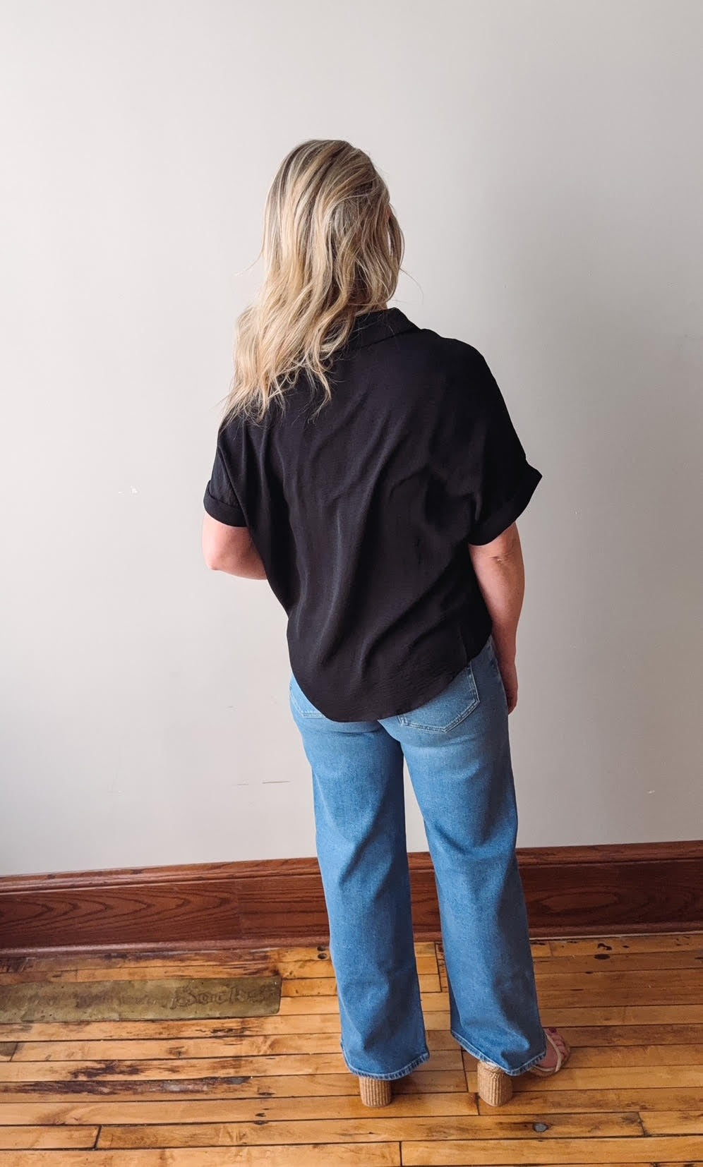 Person wearing a black shirt and blue jeans standing against a white wall on a wooden floor.