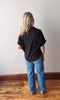 Person wearing a black shirt and blue jeans standing against a white wall on a wooden floor.