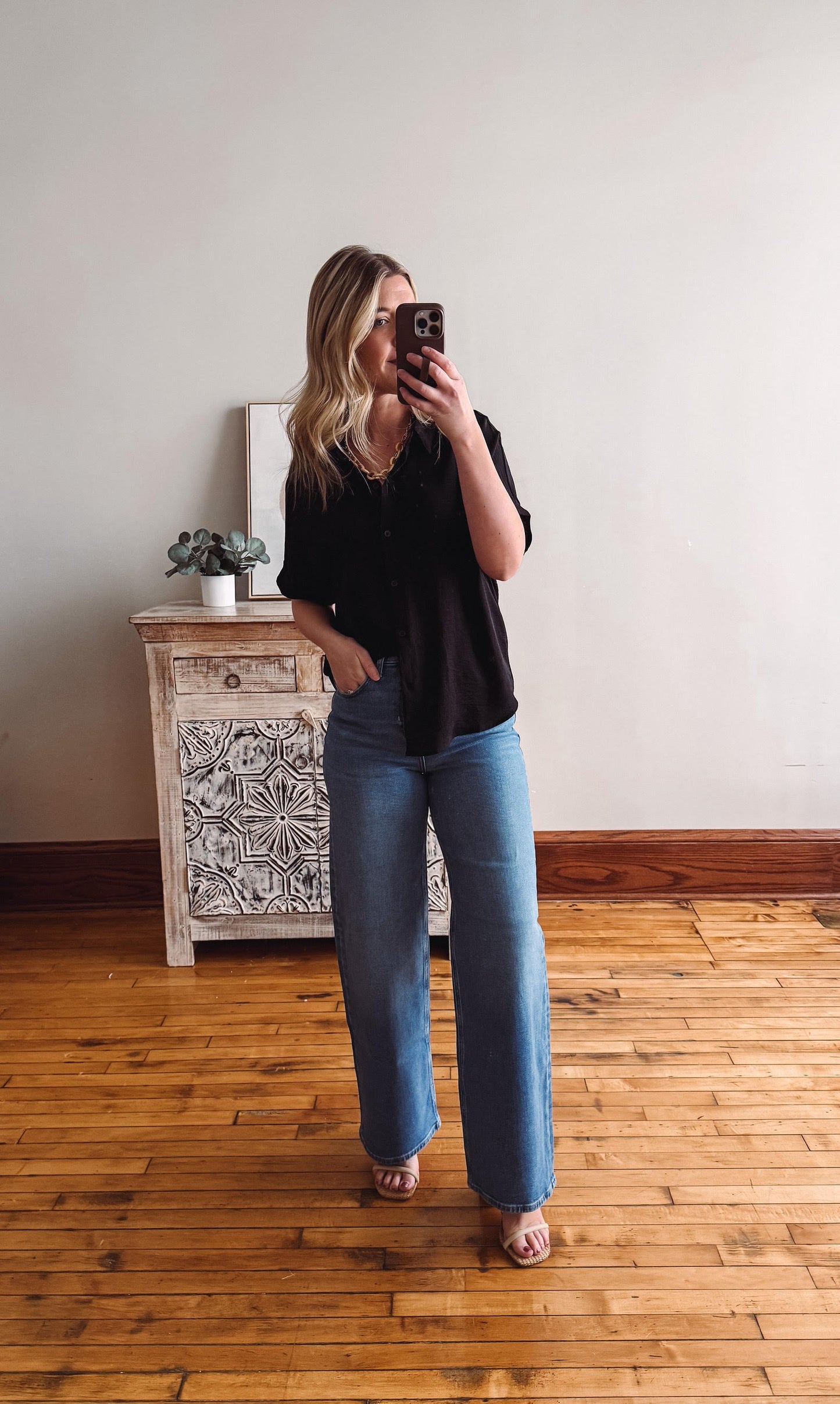Woman taking a mirror selfie wearing a black top and blue jeans in a room with wooden flooring and a decorative cabinet.