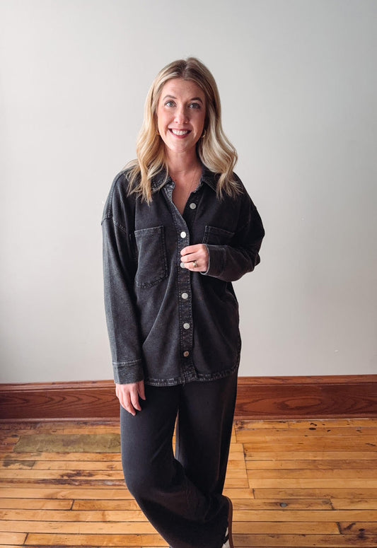 Woman wearing a dark denim jacket and black pants standing on a wooden floor with a white wall background
