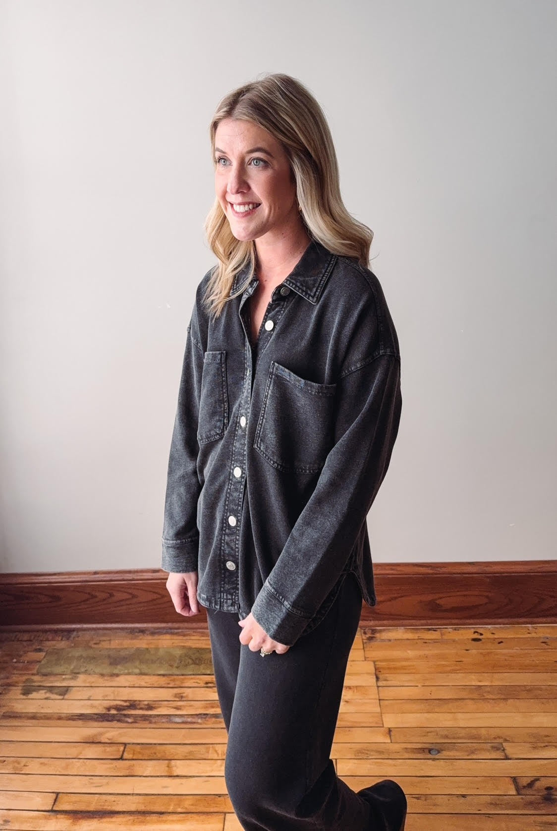 Woman wearing a dark denim jacket standing on a wooden floor with a plain wall background