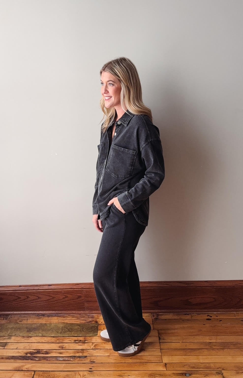 Woman wearing a dark denim jacket and black pants standing against a plain wall.