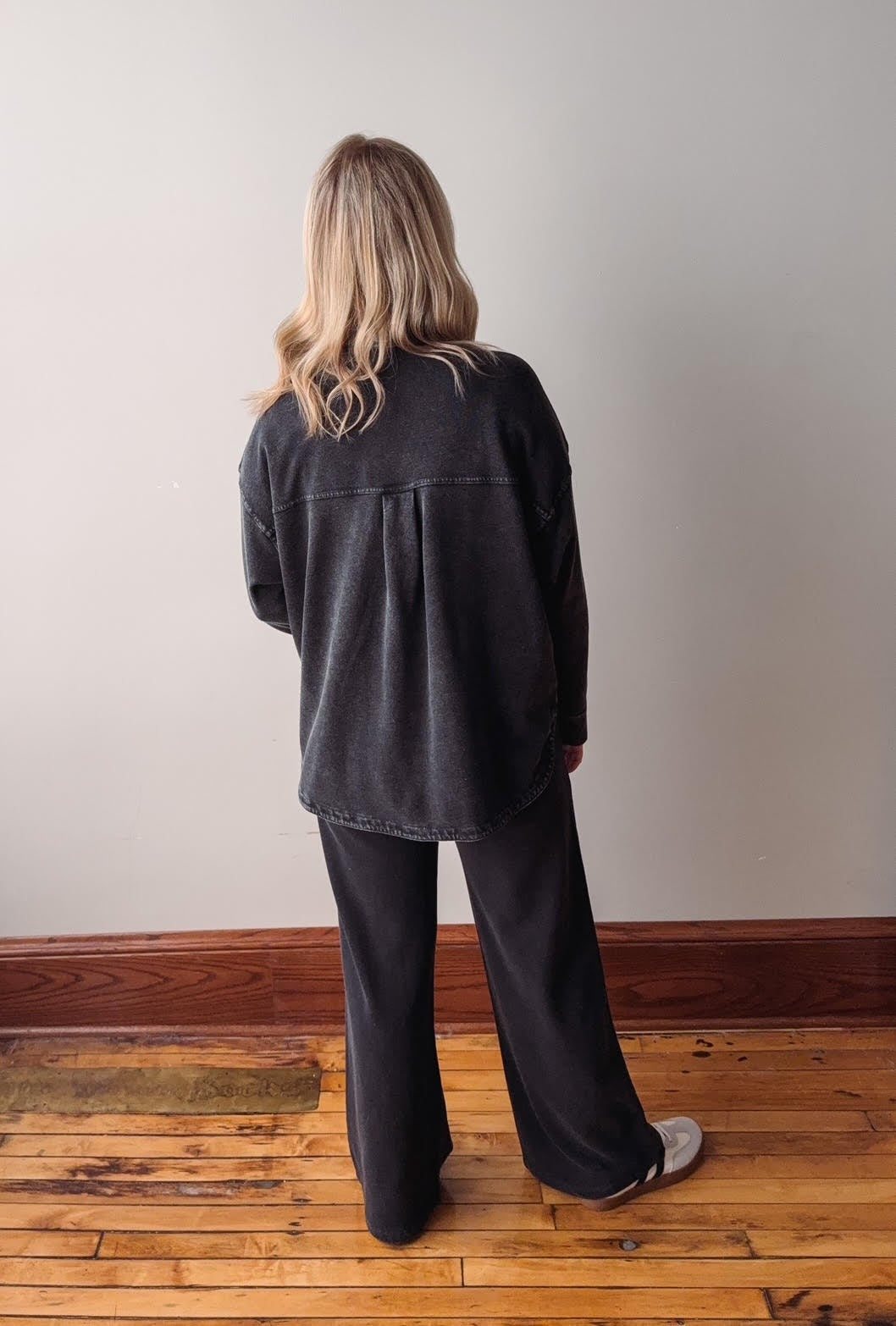 Person wearing a black jacket and pants standing on a wooden floor with a plain wall background