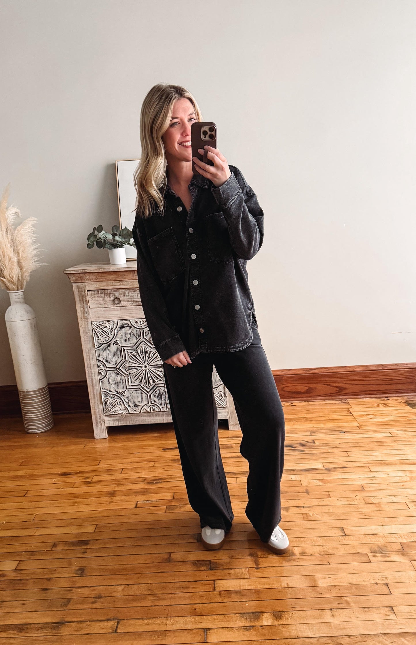 Person taking a mirror selfie wearing a dark jacket and pants in a room with wooden flooring and a decorative cabinet.