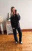 Person taking a mirror selfie wearing a dark jacket and pants in a room with wooden flooring and a decorative cabinet.