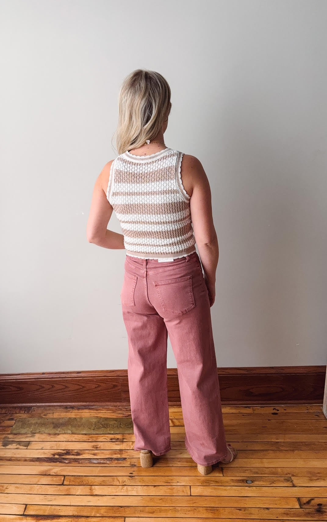 Person wearing a striped sleeveless top and pink pants standing in a room with wooden flooring and a white wall.