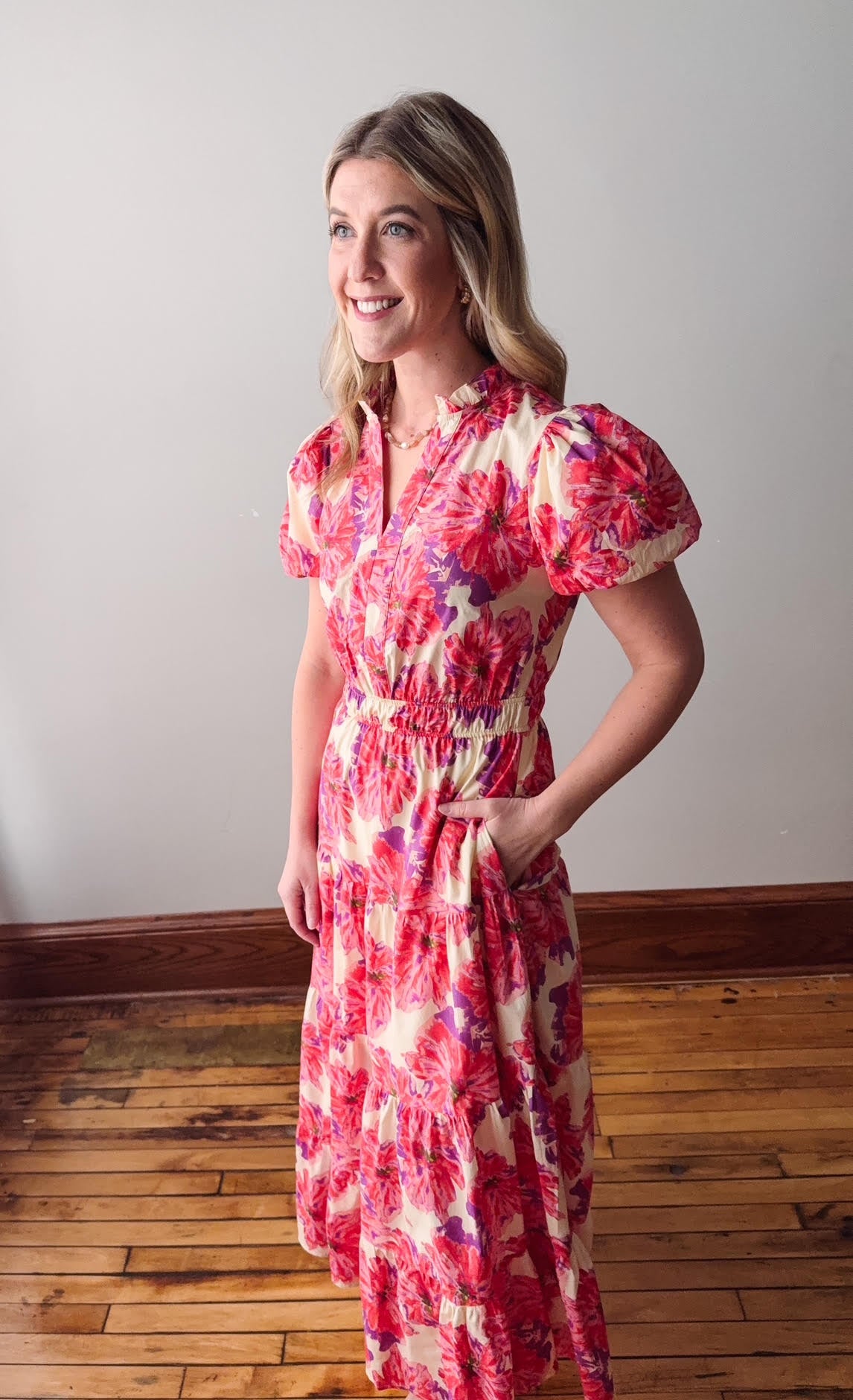 Woman wearing a red floral dress standing on a wooden floor with a white wall background