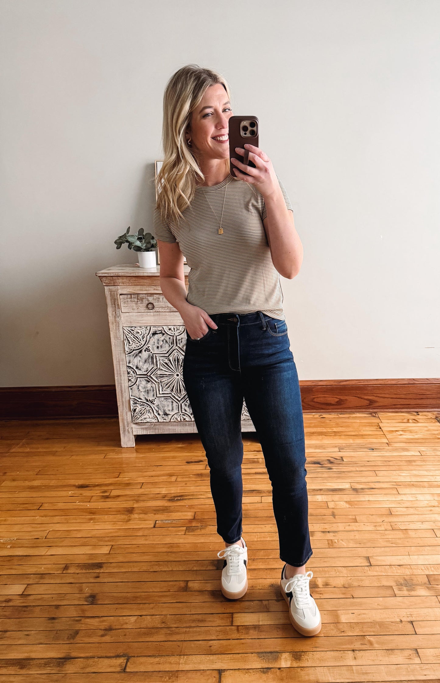 Woman taking a mirror selfie wearing a beige t-shirt and blue jeans in a room with wooden flooring.