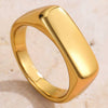 A polished yellow gold signet ring with a rounded rectangular face, resting on a light beige textured surface.