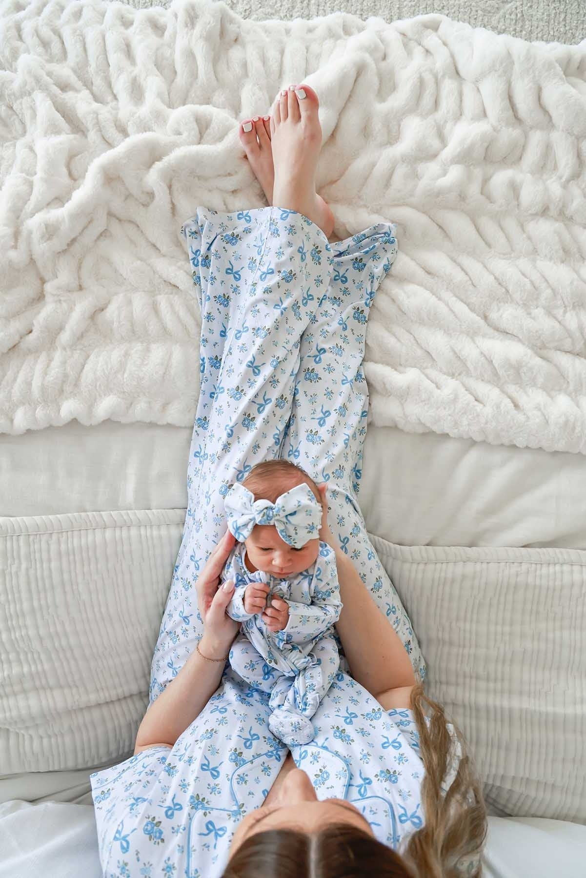 A baby wearing a matching blue and white floral onesie and bow headband lies on their back on a textured white blanket, with the mother's bare feet visible above them.