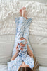 A baby wearing a matching blue and white floral onesie and bow headband lies on their back on a textured white blanket, with the mother's bare feet visible above them.